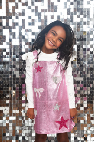MIA New York Icon Overall Dress - Light Pink
A young girl smiling in front of a silver mirror mosaic wall, wearing a shiny pink pinafore-style dress with star and heart appliqués over a white long-sleeve top.