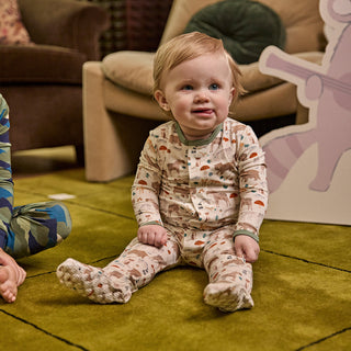 Magnetic Me Modal Magnetic Footie - Wonderful Wildlife
A baby sitting on a green carpet wearing a long-sleeve footed pajama romper with a woodland tree pattern. The background shows a cozy living room setting with a brown armchair and a large cartoon character cutout.