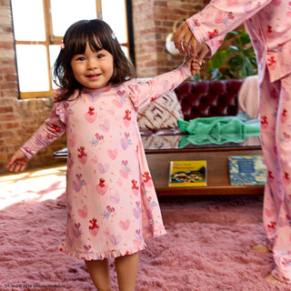 Magnetic Me x Sesame Street RightFit™ Magnetic Nightgown - Elmo & Abby
A young child wearing a pink long-sleeve pajama dress with a teddy bear and heart pattern, holding hands with an adult in matching pajamas in a cozy living room setting.