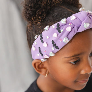 Sweet Wink Bat Halloween Gemstone Headband
A close-up of a child wearing a lavender purple knotted fabric headband decorated with black bat silhouettes, white and clear rhinestone hearts, and small iridescent gems.