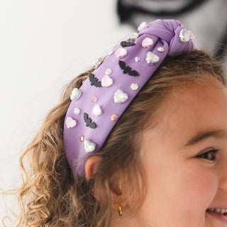 Sweet Wink Bat Halloween Gemstone Headband
A close-up lifestyle shot of a young girl wearing a purple knotted headband decorated with 3D white and pink hearts, black bat silhouettes, and small rhinestone embellishments.