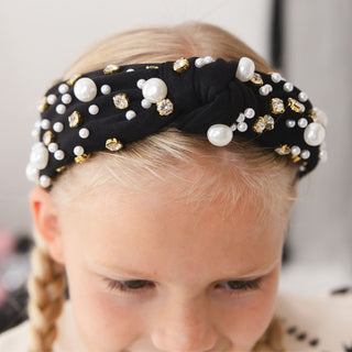 Sweet Wink Black Pearl Gemstone Headband
A close-up of a young girl wearing a black twisted fabric headband decorated with white faux pearls and gold rhinestone accents.