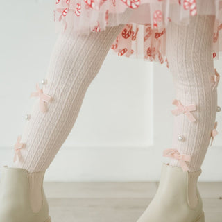 Sweet Wink Bedazzled Coquette Tights - Pink
Close-up of a person wearing light pink tights with bow details and a floral dress, standing on a light wooden floor.