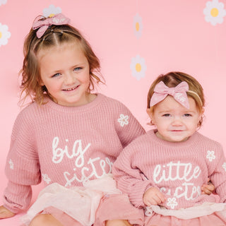 Sweet Wink Big Sister Knit Sweater
Two young girls wearing pink sweaters with 'big sister' and 'little sister' text on a pink background with flower patterns.