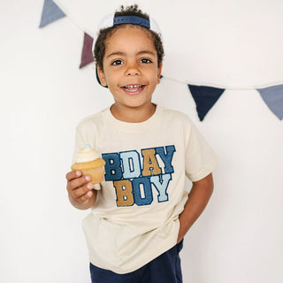 Sweet Wink Birthday Boy Patch Short Sleeve T-Shirt - Natural 
Child wearing a 'BDAY BOY' shirt holding a cupcake with a colorful flag banner in the background.