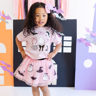 Sweet Wink Spooky Coquette Halloween Short Sleeve T-Shirt - Ballet
Young girl wearing a pink Halloween-themed dress with ghost and bat designs, standing in front of colorful decorations.
