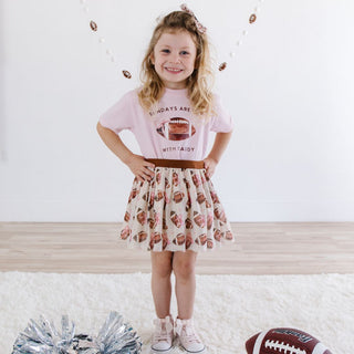 Sweet Wink Football Cutie Tutu Young girl in a pink t-shirt and patterned skirt standing on a white surface with a football and pom-poms.