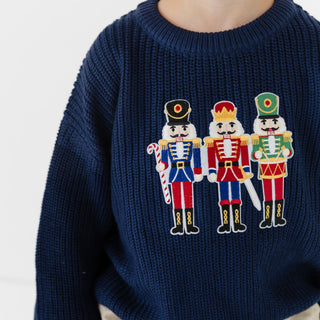 Model wearing Sweet Wink Nutcracker Patch Christmas sweater in khaki jogger style, featuring festive nutcracker patch design.