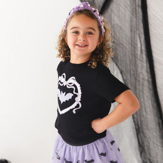 Sweet Wink Sequin Bat Halloween Tutu Skirt
A young girl with curly brown hair wearing a black short-sleeve t-shirt with a white bat graphic, a light purple glittery skirt with bat motifs, and a matching purple glittery headband, posing against a white and black backdrop.