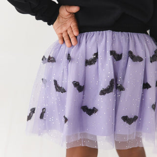 Sweet Wink Sequin Bat Halloween Tutu Skirt
A close-up of a child wearing a lavender tulle tutu skirt with black bat appliqués and white sequins, paired with a black top, against a plain light background.