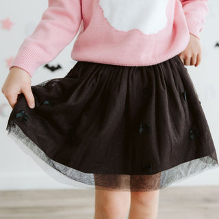 Sweet Wink Onyx Bow Tutu Skirt
A child wearing a pink sweater with a white cloud detail and a black tulle skirt decorated with small black bat shapes, shown from the chest down in a bright indoor setting.
