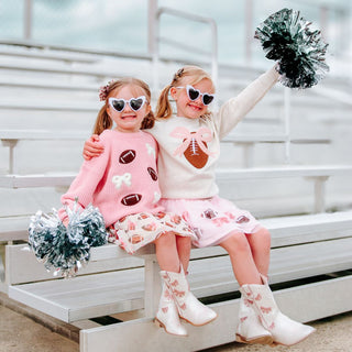 The Coquette Football Sequin Tutu is a fun skirt and dress up tutu for celebrating Game Day!

Features:

Ballet pink tulle and cotton lining
Brown and white sequin footballs and pink sequin bows
Pink glitter elastic waistband
Care: Hand wash and line dry
The Sweet Wink difference:

Super soft tulle that doesn’t scratch or itch
Cotton lining provides extra coverage and comfort
“Shed-free” embellishments ensure glitter does not sprinkle arouTwo young girls in matching outfits with pom-poms sitting on a bench.