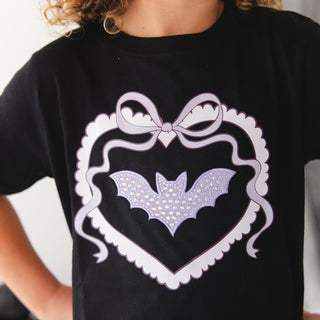 Sweet Wink Rhinestone Bat Patch Halloween Short Sleeve T-Shirt
A close-up of a child wearing a black short-sleeve T-shirt with a large white heart outline, bow, and bat graphic on the front. The bat is decorated with small rhinestones.