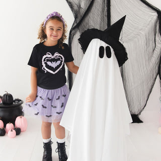Sweet Wink Rhinestone Bat Patch Halloween Short Sleeve T-Shirt
A young girl stands next to a large ghost costume prop in a Halloween-themed studio setup. The girl wears a black graphic T-shirt with a bat-heart design, a light purple tutu skirt with black bat prints, white socks, and black boots.