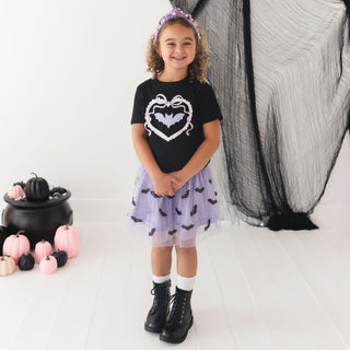 Sweet Wink Rhinestone Bat Patch Halloween Short Sleeve T-Shirt
A young girl stands in a studio wearing a black short-sleeve T-shirt with a white and purple bat-heart graphic, a light purple tutu skirt with black bat prints, white socks, and black boots. She has a purple bat headband.