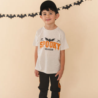 Sweet Wink Spooky Season Halloween Short Sleeve T-Shirt
A young boy wearing a light gray heather t-shirt with a 'Spooky Season' graphic in orange and black, paired with black pants and a black baseball cap, standing against a beige wall with a bat banner.