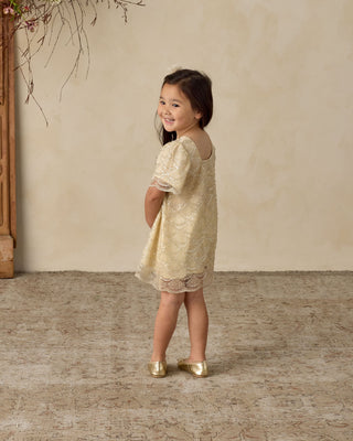 Noralee Daisy Dress - Silver Metallic
Young girl in a gold lace dress standing against a beige wall.