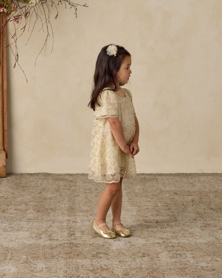 Noralee Daisy Dress - Silver Metallic
Young girl in a lace dress standing against a beige wall.