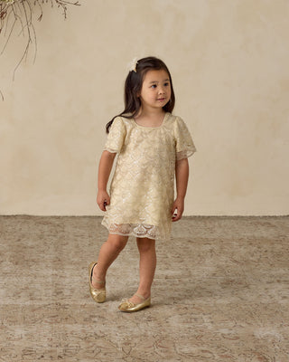 Noralee Daisy Dress - Silver Metallic
Young girl in a beige lace dress standing on a textured surface with a neutral background