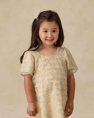 Noralee Daisy Dress - Silver Metallic
Young girl wearing a gold sequin dress against a beige background