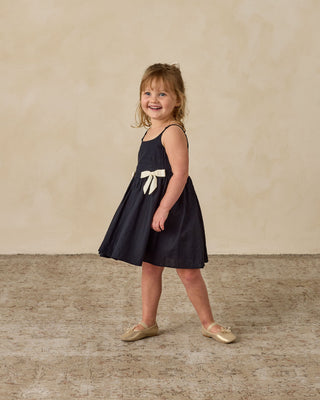 Noralee Betsy Dress - Black
Young girl wearing a navy dress with a white bow, standing on a textured floor against a beige wall.
