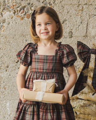 Noralee Louella Dress - Winter Tartan
Young girl in a plaid dress holding a wrapped gift against a stone wall.