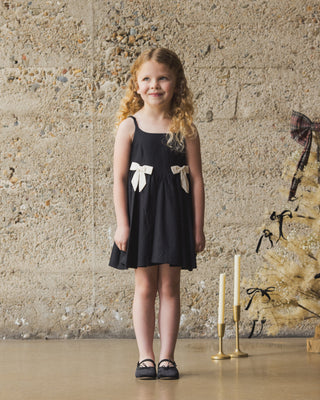 Noralee Betsy Dress - Black
Young girl in a black dress with white bows standing against a textured wall.