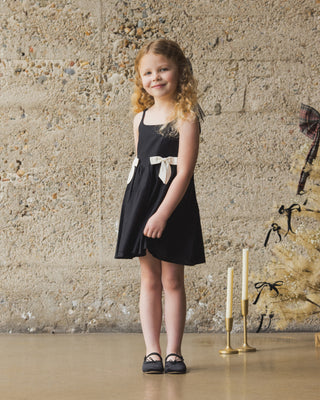 Noralee Betsy Dress - Black
Young girl in a black dress standing in front of a textured wall with candles and decorative elements.