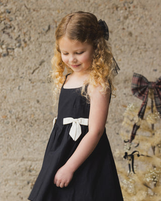 Noralee Betsy Dress - Black
Young girl in a black dress with a white bow standing against a textured wall.