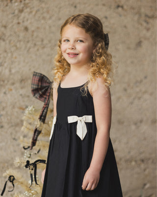 Noralee Betsy Dress - Black
Young girl in a black dress with a white bow standing against a textured wall.