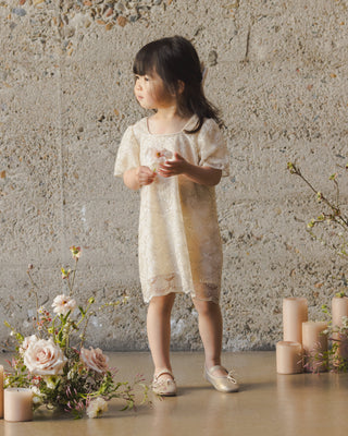 Noralee Daisy Dress - Silver Metallic
Young girl in a light dress standing in a room with flowers and candles.
