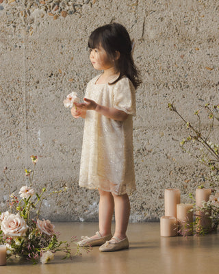 Noralee Daisy Dress - Silver Metallic
Young girl in a beige dress standing in a room with flowers and candles.