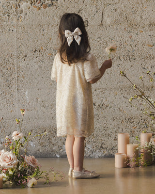 Noralee Daisy Dress - Silver Metallic
Young girl in a floral dress holding a flower, standing against a textured wall with flowers and candles on the floor.
