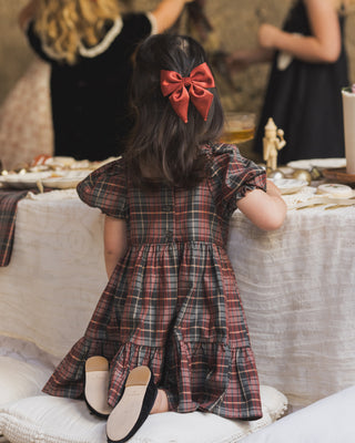 Noralee Louella Dress - Winter Tartan
Child in a plaid dress with a red bow sitting at a table with people in the background.