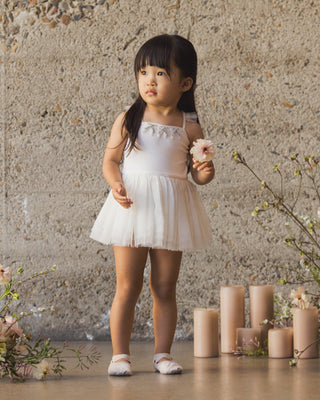 Noralee Clementine Tutu - Ivory
Young girl in a white dress standing in front of a stone wall with candles and flowers.