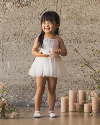 Noralee Clementine Tutu - Ivory
Young girl in a white dress standing in front of a stone wall with candles and flowers.