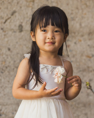Noralee Clementine Tutu - Ivory
Young girl in a white dress holding a flower against a textured wall.