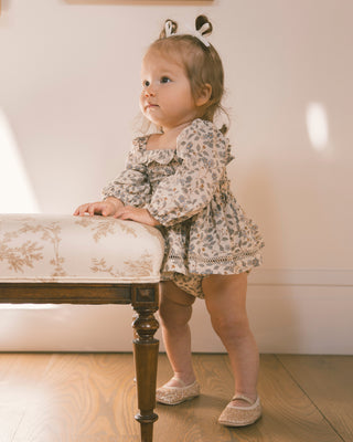 Noralee Elodie Romper - Blue Garden
A toddler girl wearing a floral long-sleeve romper and matching diaper cover stands on a wooden floor next to a bench, looking off-camera. She has a hair bow accessory on her head.
