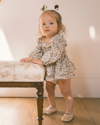 Noralee Elodie Romper - Blue Garden
A toddler girl wearing a beige and brown floral print long-sleeve romper dress and matching pointed shoes, standing next to a wooden bench with a floral upholstered seat.