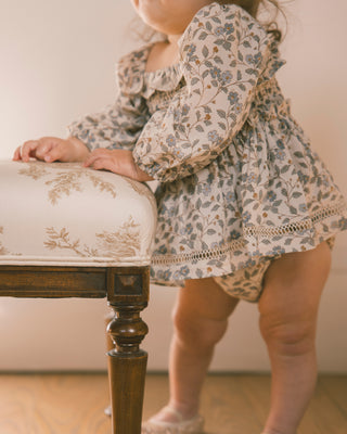 Noralee Elodie Romper - Blue Garden
A toddler wearing a long-sleeve floral dress with puff sleeves and a ruffled neckline, standing beside a wooden bench with cream floral upholstery in a softly lit indoor setting.