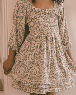 Noralee Elodie Dress - Blue Garden
A woman wearing a cream-colored smocked dress with a blue and brown floral print, featuring ruffled trim and elbow-length sleeves.