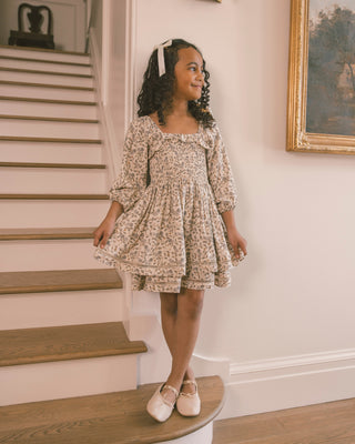 Noralee Elodie Dress - Blue Garden
A young girl with curly dark hair wearing a beige and brown floral print dress, standing on a wooden staircase in a home interior.