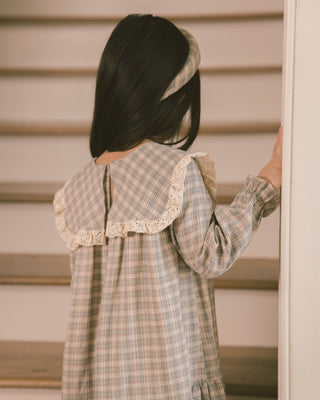 Noralee Noelle Dress - Fall Plaid
A child wearing a beige and gray plaid long-sleeve dress with a large ruffled collar and lace trim, shown from behind in an indoor setting with wooden steps.