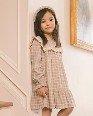 Noralee Noelle Dress - Fall Plaid
A young girl wearing a beige and brown plaid dress with ruffle details and a matching headband, standing in an indoor setting with a cream-colored wall and molding.