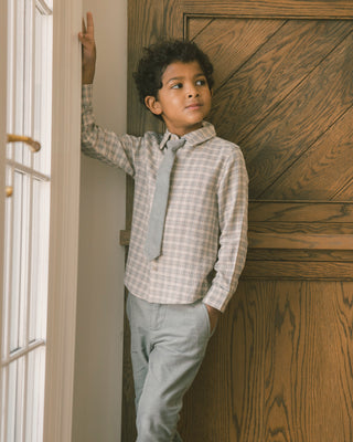 Noralee Harrison Button Down - Fall Plaid
A young boy wearing a light beige and white plaid button-down shirt, a matching light gray tie, and gray trousers, leaning against a white door frame and a wooden paneled wall.