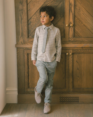 Noralee Harrison Button Down - Fall Plaid
A young boy wearing a light beige and white checkered shirt, a matching light gray tie, light blue jeans, and beige sneakers, standing in front of a large wooden cabinet.
