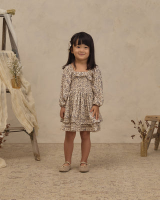 Noralee Elodie Dress - Blue Garden
A young girl with dark hair stands in a studio setting wearing a beige and brown floral print long-sleeve dress with a smocked bodice, paired with beige ballet flats.