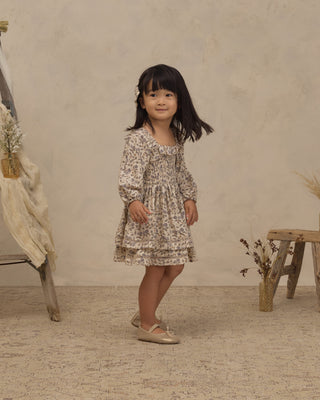 Noralee Elodie Dress - Blue Garden
A young girl with dark hair stands in a studio setting wearing a long-sleeve, square-neck babydoll dress with a delicate beige and brown floral pattern. The background features a textured beige wall, a rustic wooden stool with dried flowers, and a draped fabric on a wooden stand.