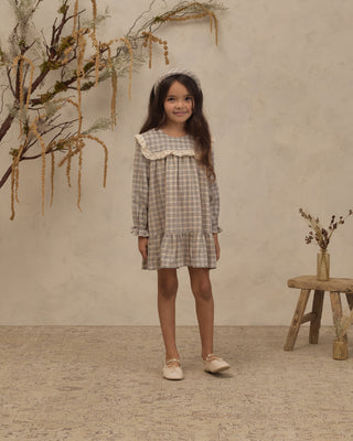 Noralee Noelle Dress - Fall Plaid
A young girl stands in a studio setting wearing a long-sleeve plaid dress with a ruffled collar and a matching headband. The background features a textured beige wall, dried botanical decorations, and a small wooden stool.