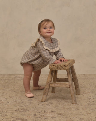 Noralee Noelle Romper - Fall Plaid
A toddler girl wearing a beige and brown plaid romper with a large cream lace collar stands on a rustic wooden stool against a textured beige backdrop.